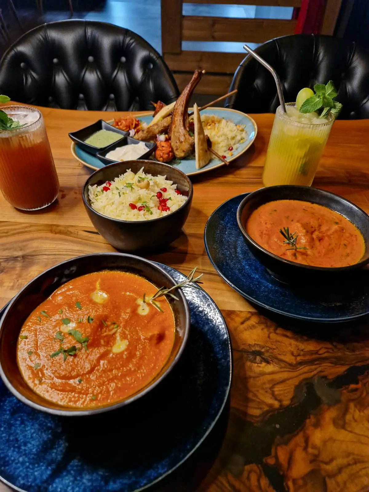 Gedeckter Holztisch bei Kahani: zwei Curries in blauen Schalen, Reis, Grillteller und Limonaden